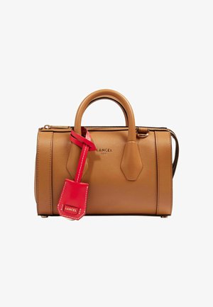 Sac à main en cuir fauve avec double poignées et une étiquette en cuir rouge attachée, portant le nom de la marque Lancel à la fois sur le sac et l'étiquette.