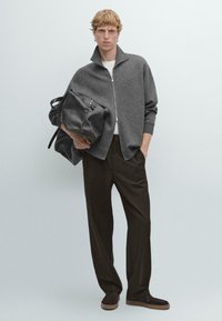 Maglione grigio con zip, camicia bianca, pantaloni larghi marroni, tiene una borsa in pelle nera. Texture morbida, design casual, palette di colori neutri, accenti minimali.