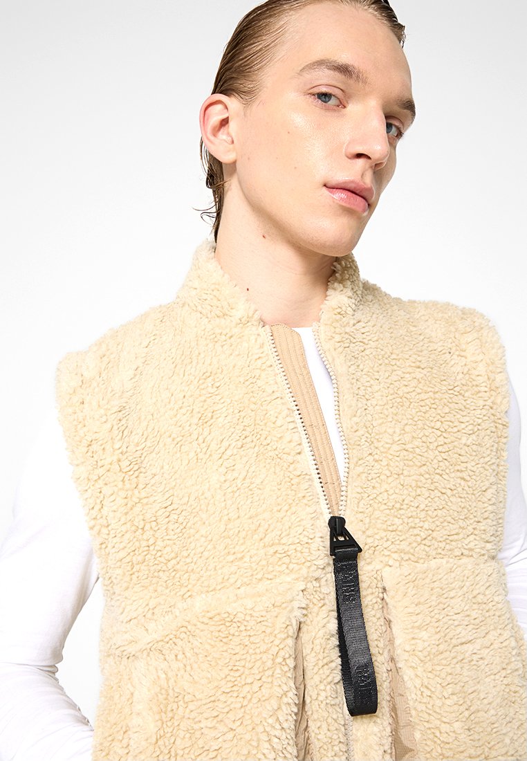 Gilet in pile beige con zip frontale, due tasche laterali e una superficie testurizzata. Il gilet presenta un rivestimento interno a contrasto color nocciola e una zip nera.
