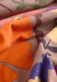 Gros plan d’un tissu doux avec des motifs floraux colorés, incluant de grands pétales orange, violets et verts sur un fond rose atténué.