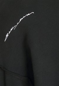 Tessuto nero con testo corsivo bianco "Blumarine" ricamato vicino a una cucitura piegata.