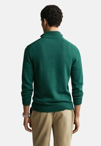 Grüner gestrickter Pullover mit hohem Kragen, mit gerippten Bündchen und Saum, von hinten gesehen, getragen mit hellen Hosen.