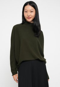 Giovane donna con lunghi capelli neri che indossa un maglione largo verde scuro e una gonna plissettata nera, in piedi davanti a uno sfondo bianco.