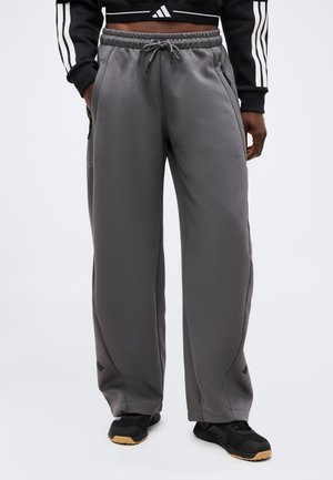 Partie médiane montrant une personne portant un pantalon de survêtement gris ample avec cordon et baskets noires, mains dans les poches, haut de sport noir court avec des bandes blanches.