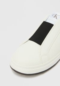 Λευκό αθλητικό παπούτσι χωρίς κορδόνια με μαύρη ελαστική ταινία στο επάνω μέρος, ομαλή υφή, στρογγυλεμένη μύτη και λαστιχένια σόλα. Διακριτικές ραφές.