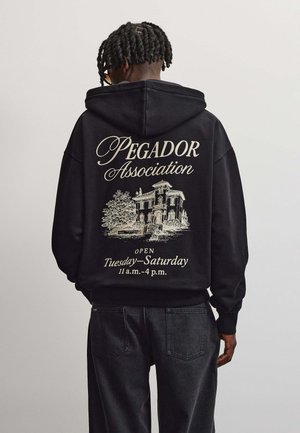 Person wearing a black hoodie with "Pegador Association" text and a house graphic, open Tuesday to Saturday 11 a.m. to 4 p.m., facing away.