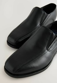 Zapatos negros sin cordones, hechos de cuero suave, con una punta redondeada, detalles cosidos y lados elásticos para facilitar su uso.