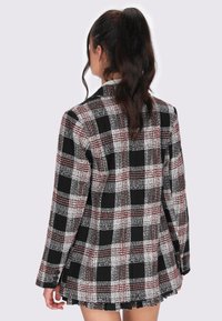 Donna con capelli scuri raccolti in un alto ponytail, che indossa un blazer a quadri nero, bianco e rosso con una gonna abbinata, vista da dietro.