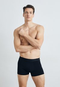 Černé boxerky vyrobené z hladké tkaniny, s širokým elastickým páskem se značkou, nošené bez trička mužským modelem.