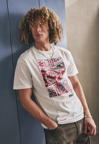 Valge puuvillane T-särk, millel on punane ja must graafiline print, abstraktne tekst ja kujundid, ümmargune kaelus ja lühikesed varrukad.