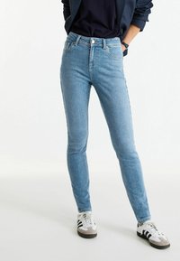 Jeans de tiro alto en azul claro con corte ajustado, que cuentan con bolsillos frontales y cierre de botón. Combinados con zapatillas blancas y grises.