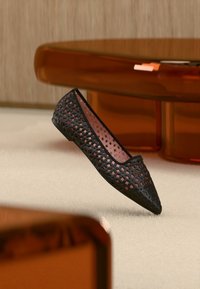 Μαύρο γυναικείο παπούτσι με μυτερή μύτη και επίπεδη σόλα, με γεωμετρικό μοτίβο κοπής, τοποθετημένο σε ανοιχτόχρωμο χαλί κοντά σε διαφανή πορτοκαλί έπιπλα.