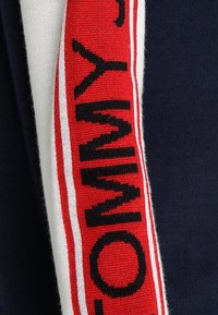 Pull bleu marine avec un panneau vertical rouge et blanc affichant "TOMMY" en lettres noires. Texture en maille douce avec des détails de rayures contrastantes.