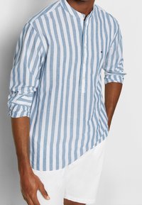 Camicia in lino a righe blu e bianche con colletto a camicia, apertura con bottoni e maniche lunghe, indossata con pantaloni corti bianchi. Logo discreto sul petto.