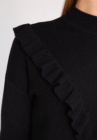 Svart ribbat stickat tröja med en bred volangdetalj längs ena axeln. Hög hals med slät textur och åtsittande silhuett.