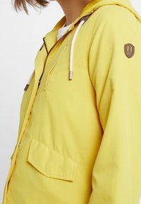 Gul, letvægtsjakke med hætte. Har en forlommer, snoretræk og et mærke på skulderen. Glat tekstur.