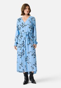 Light blue long-sleeve dress with a v-neck, black leaf pattern, button details, waist tie, and gathered sleeves, worn with black ankle boots.