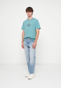 Šviesiai mėlynos džinsinės kelnės derinamos su turkio spalvos medvilniniu marškinėliu, kuriame centre yra tamsus logotipas. Modelis dėvi baltus sportinius batelius, stovėdamas prieš paprastą foną.