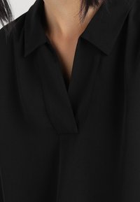 Černá blusa s límečkem, výstřihem do V a hladkým textilním povrchem. Detailní záběr zdůrazňuje čisté linie a minimalistický design.