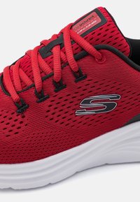 Chaussure de sport rouge avec une tige en mesh respirant, accents noirs et semelle blanche rembourrée. Présente un logo bien visible et des lacets texturés.