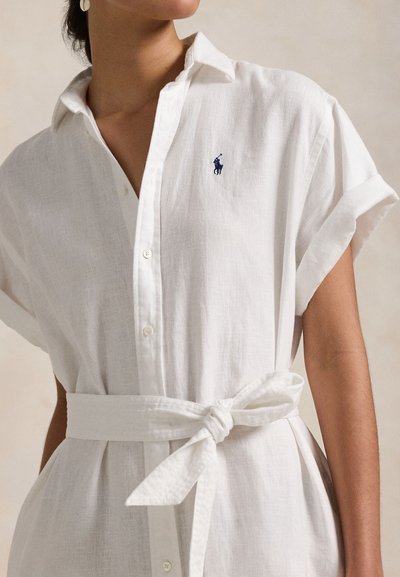 Femeie purtând o rochie cămașă din in alb cu curea, mâneci suflecate și un mic logo polo jucător în albastru închis pe piept.