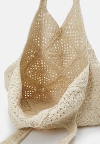 Crochetad beige tote väska med en öppen, diamantmönstrad design. Texturerat tyg och två robusta axelremmar. Rymligt inre.