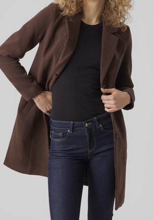 Femme portant un manteau brun foncé sur un haut noir et un jean bleu foncé, se tenant debout les mains ajustant le devant du manteau.