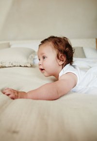 Bebeluș într-un costum alb fără mâneci, târându-se pe o suprafață bej texturată înconjurată de perne moi cu margini uzate. Ochii albaștri vizibili.