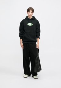 Hanorac negru cu logo oval verde, buzunar kangaroo; asortat cu pantaloni negri largi și pantofi albi; purtând o geantă neagră.