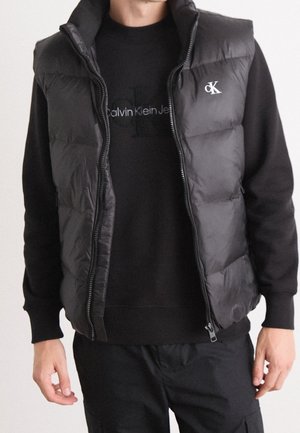 Homme portant un sweat-shirt Calvin Klein Jeans noir et un gilet matelassé noir avec logo CK, montré du cou aux hanches sur fond uni.