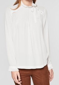 Weiße Bluse mit vertikalen Falten, hohem Ausschnitt mit Bindeschluss und langen Ärmeln mit Rüschendetails. Glattes Material, lockere Passform.