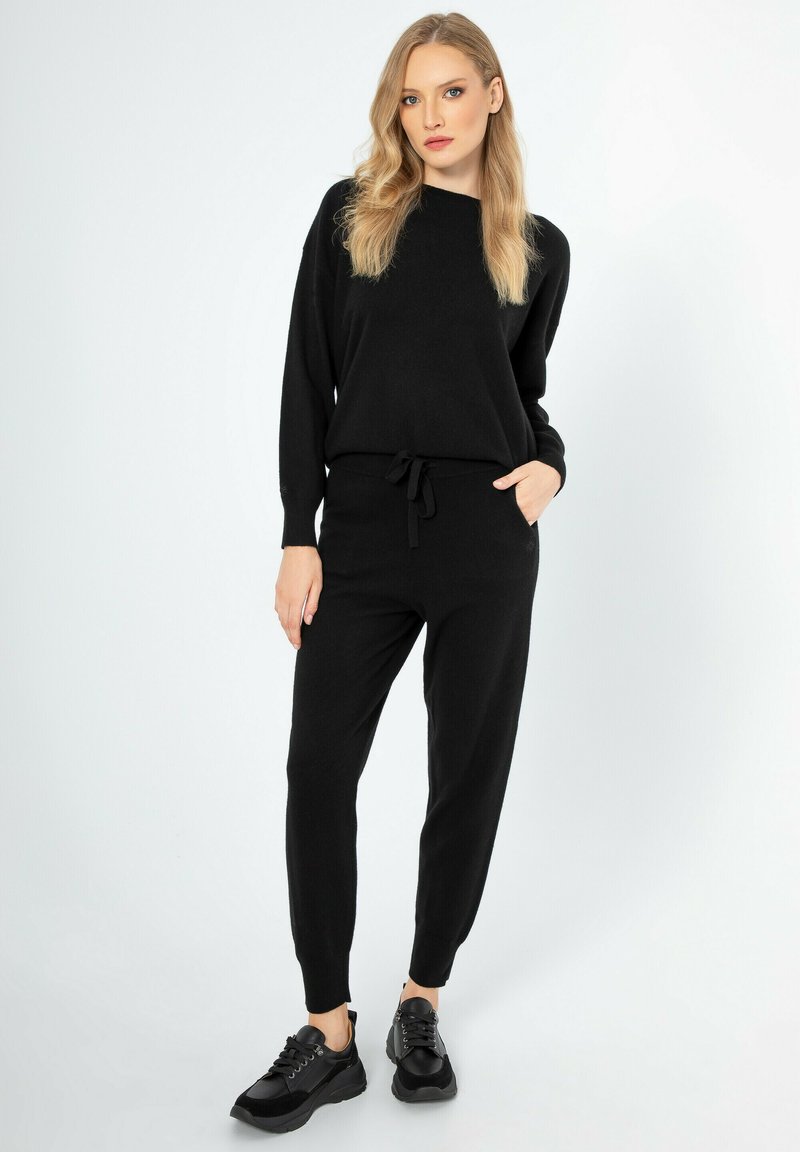 Schwarzer gestrickter Jumpsuit mit langen Ärmeln, schmalen Bündchen und einem Kordelzug in der Taille. Kombiniert mit schwarzen Schnürsenkelschuhen. Einfaches, nahtloses Design.