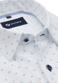 Witte blouse met een blauw micro-patroon, contrasterende blauwe kraagvoering en marineblauwe knopen met een label met de tekst "SUITABLE."