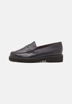 Zwarte leren loafer met dikke geribbelde zool en decoratief banddetail aan de bovenkant, weergegeven op een witte achtergrond.