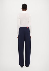 Navy wide-leg trousers with a smooth texture and high waistband. Features a red label detail at the back, paired with a white long-sleeve top.