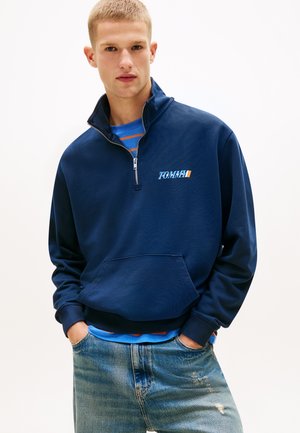 Bărbat tânăr cu păr scurt, blond, purtând un hanorac bleumarin cu fermoar scurt, cămașă albastră în dungi dedesubt și blugi decolorați, cu mâinile în buzunare.