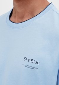Himmelblå sweatshirt med rund hals og mørkeblå kant, der har en trykt etikette med teksten "Himmelblå" nær kraven.