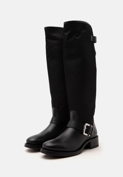 Paire de bottes hautes en cuir noir avec bouts arrondis et détail de boucle argentée sur les côtés extérieurs, sur un fond clair uni.