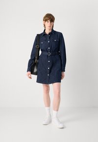 Denim shirt dress in dark blue with long sleeves, front buttons, two chest pockets, a belted waist, and white sneakers.