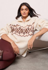 Femeie cu păr închis la culoare, purtând un pulover crem din fleece cu un model geometric, pantaloni burdundii și botine maro, așezată pe podea lângă un scaun texturat.