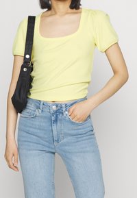 Keltainen ribbipintainen lyhythihainen paita, jossa on neliön muotoinen pääntie, yhdistettynä vaaleansinisiin korkeavyötäröisiin farkkuihin ja mustaan olkalaukkuun.