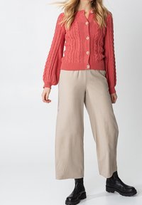 Koralstickad cardigan med kabelmönster och träknappar, i kombination med beige vida byxor och svarta robusta ankelstövlar.