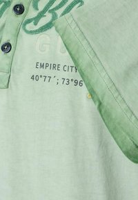 Lichtgroene polo met donkere groene accenten, drie knopen en geborduurde tekst waaronder "Empire City" en coördinaten. Zachte textuur.