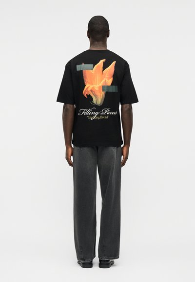 Άνδρας με την πλάτη γυρισμένη, φοράει μαύρο T-shirt με μεγάλο πορτοκαλί λουλούδι και το κείμενο "Filling Pieces 'Breaking Bread'" και χαλαρά σκούρα γκρι παντελόνια.