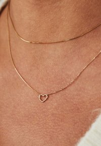 Gold chain necklace featuring a delicate heart-shaped pendant, set against skin with a subtle texture and a second thin chain visible above.