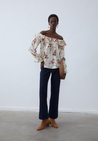 Blouse florale crème à épaules dénudées avec des bords froncés, associée à un jeans large bleu foncé et des bottines à talons marron.
