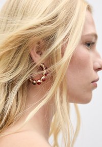 Goldener Creolenohrring mit roten und klaren Perlenakzenten. Strukturierte Oberfläche, kreisförmiges Design, getragen im Seitenprofil mit lockerem blondem Haar.