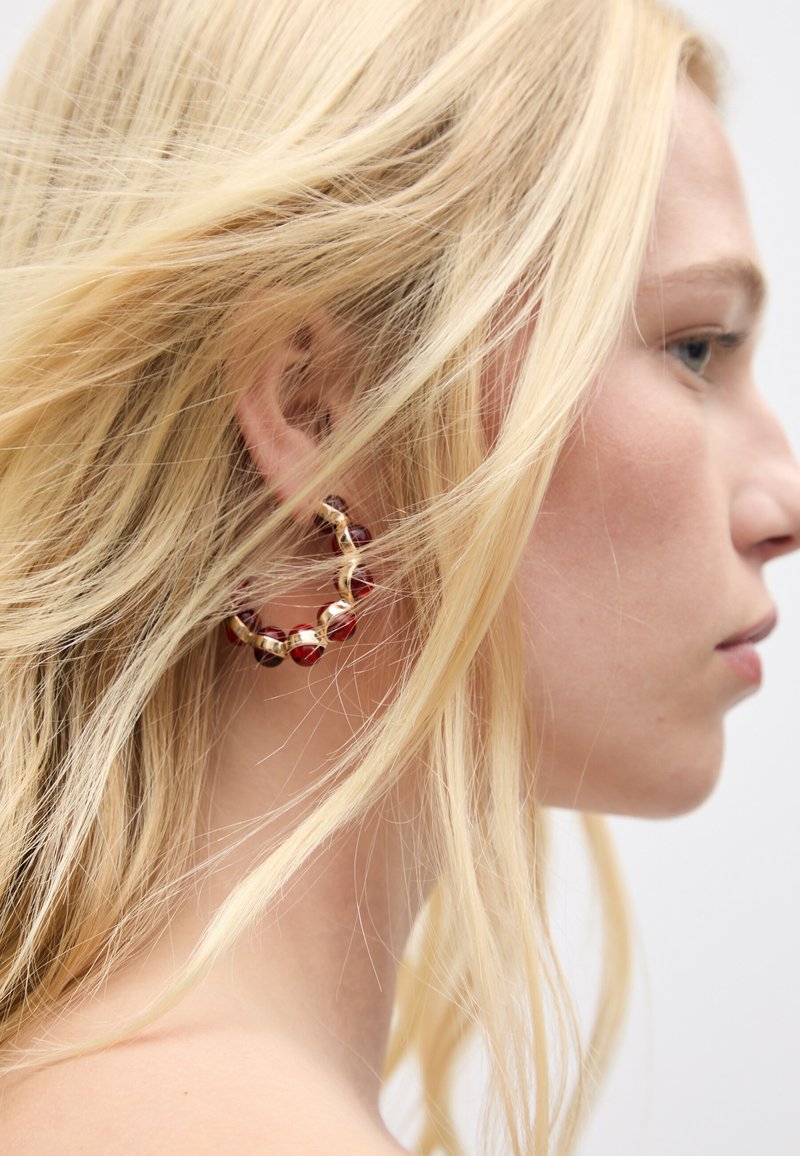Goldener Creolenohrring mit roten und klaren Perlenakzenten. Strukturierte Oberfläche, kreisförmiges Design, getragen im Seitenprofil mit lockerem blondem Haar.