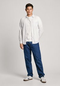 Camisa de rayas blanca de manga larga sobre una camiseta blanca, combinada con jeans de mezclilla azul y zapatillas blancas con acentos negros.