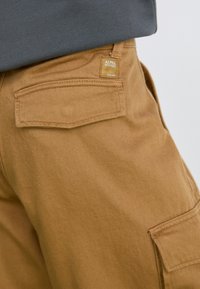 Brown cargo pants made of cotton, featuring a back pocket, a textured finish, and a small gold tag with the brand name "ALPHA INDUSTRIES."